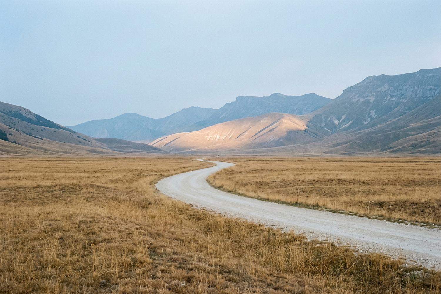 A pale track curving through dry grass toward layered ridges in morning haze.