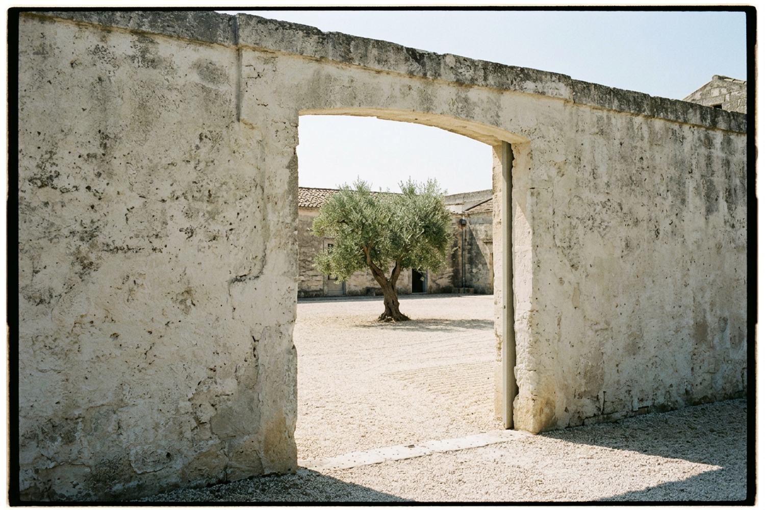 An open gateway through a thick wall onto a bright empty gravel courtyard.