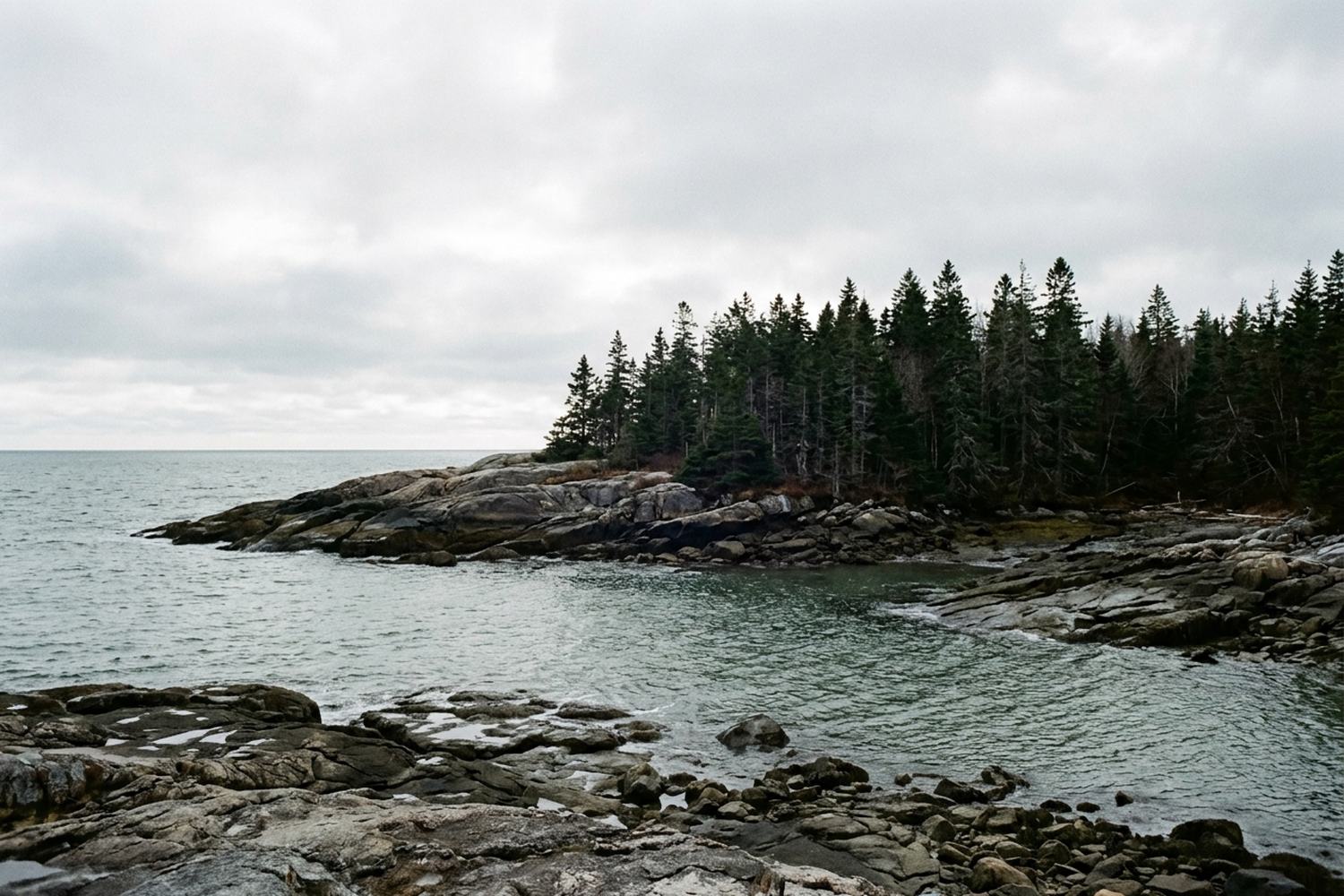 A granite headland and a narrow tidal inlet under a wide overcast sky.