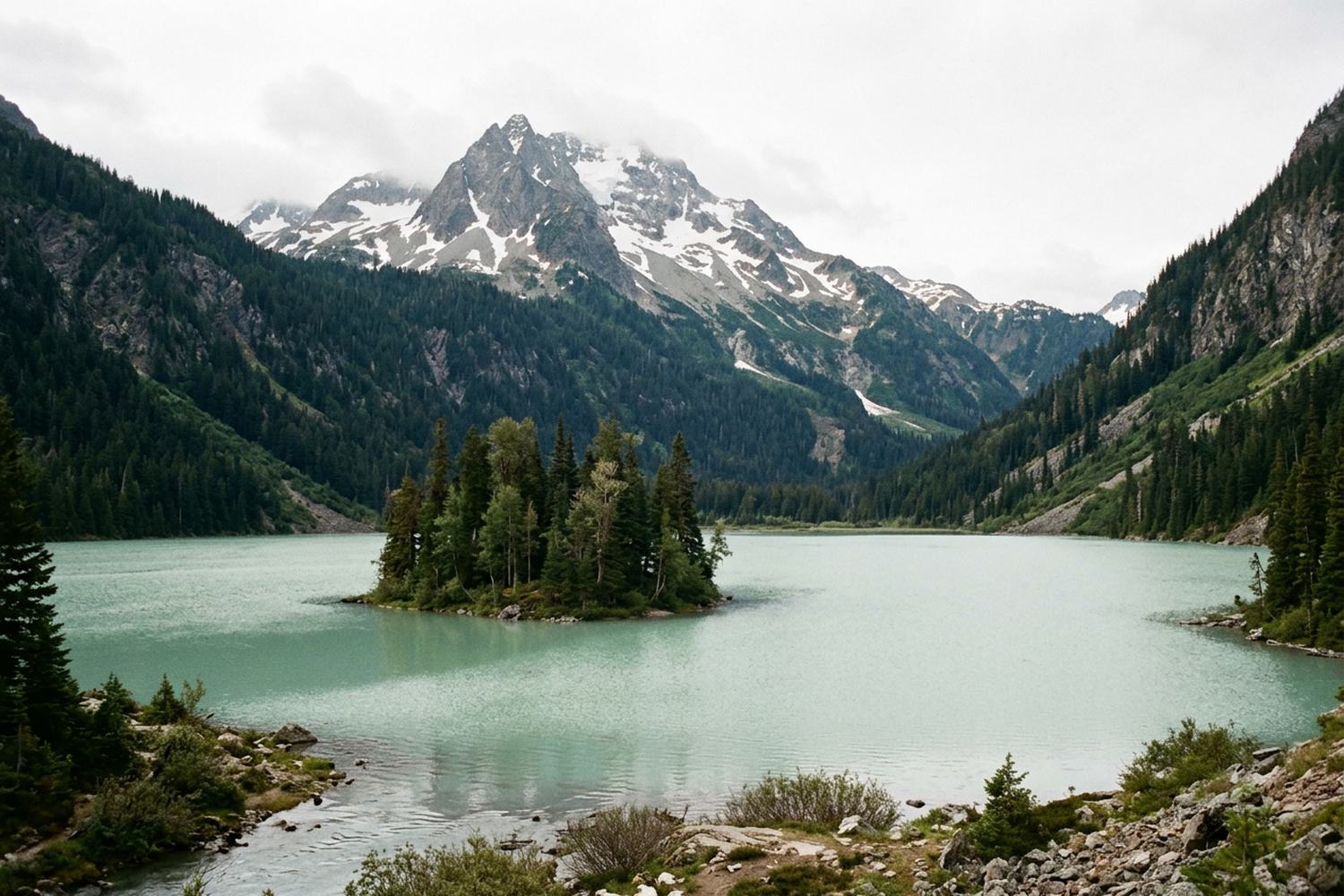 A clear alpine lake with a small wooded island below forested slopes.