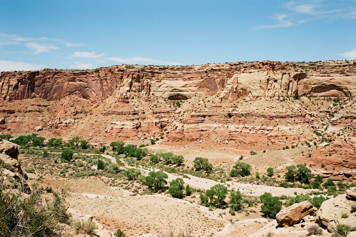 Layered red and cream sandstone cliffs above a dry canyon floor of pale sand.